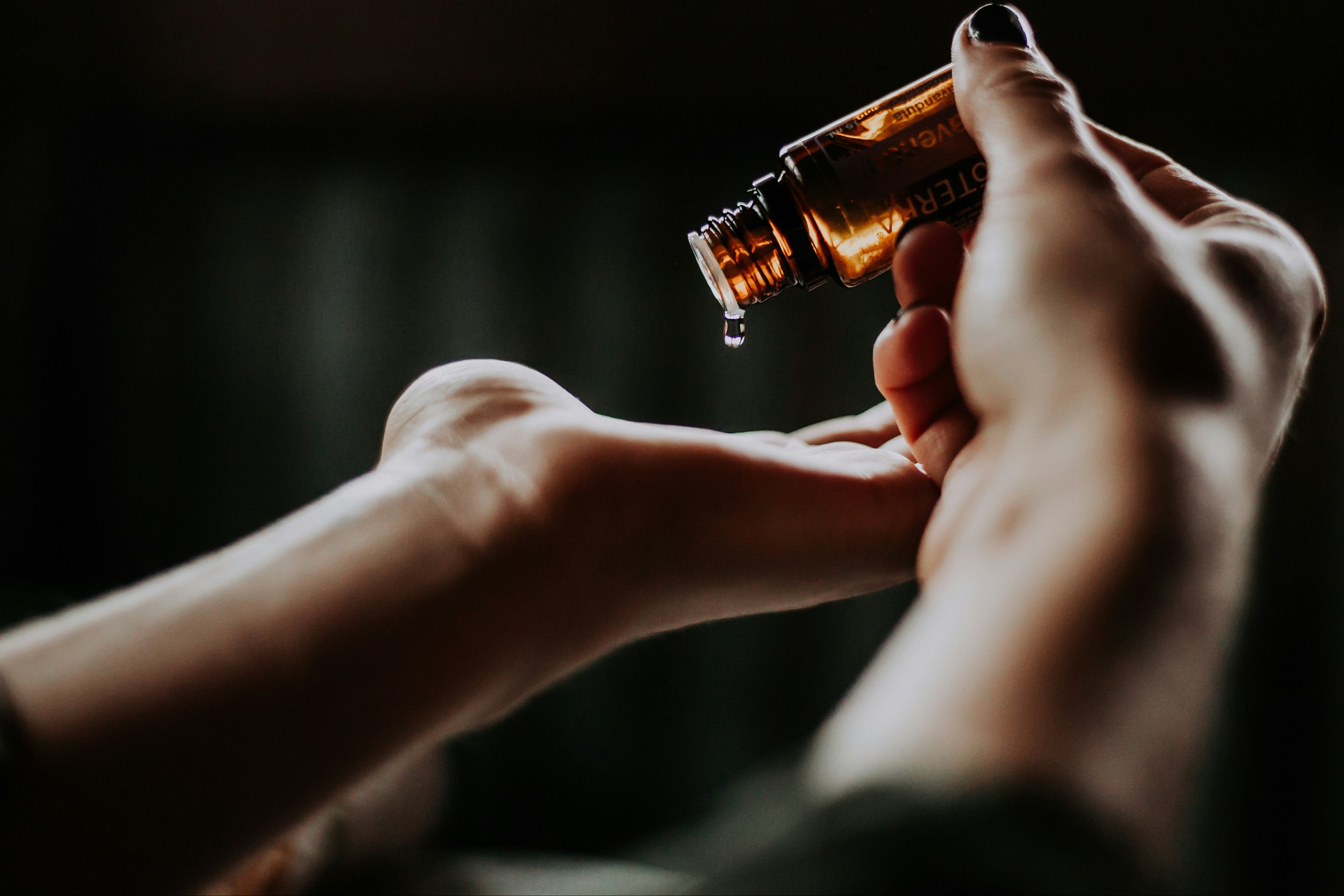 Hand holding a small bottle with a dropper over another bottle, against a dark background